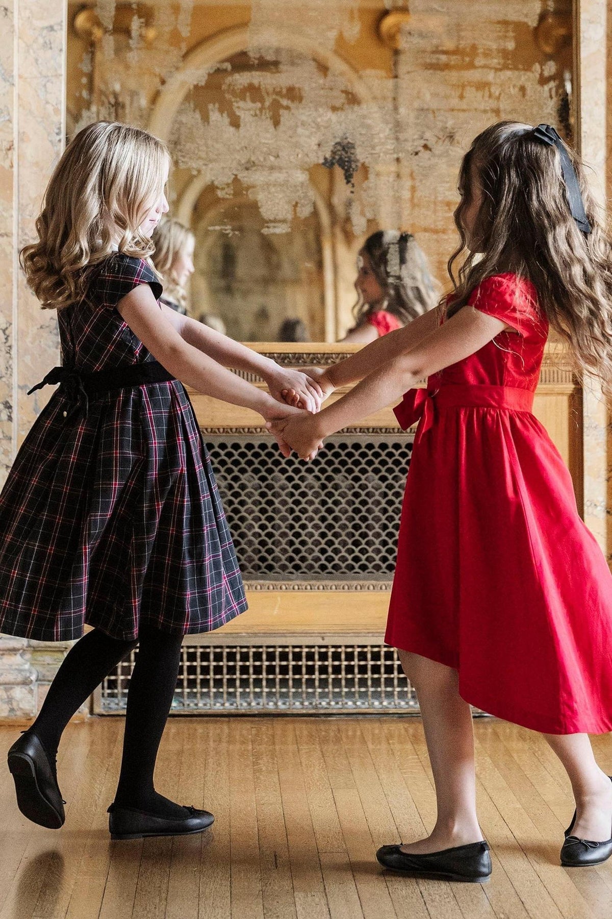 Two girls hold hands, one in a black plaid dress with cap sleeves, and the other in a red high-low.