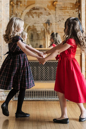 Two girls hold hands, one in a black plaid dress with cap sleeves, and the other in a red high-low.