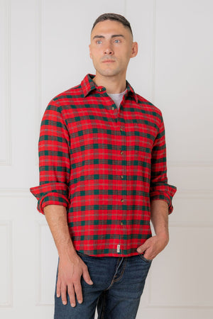 A man is wearing a red and green plaid flannel shirt, featuring a button-down collar and rolled sleeves, set against a minimalistic white wall.