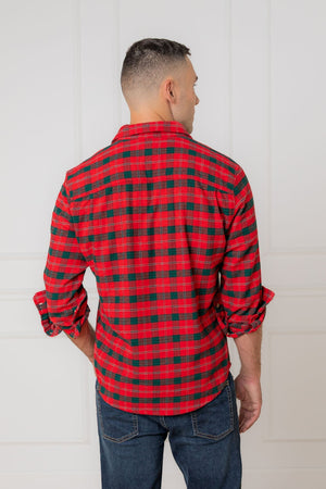 A man is seen from the back, wearing a red and green plaid flannel shirt made of organic cotton. He stands against a neutral-colored wall, showcasing the shirts vibrant pattern.