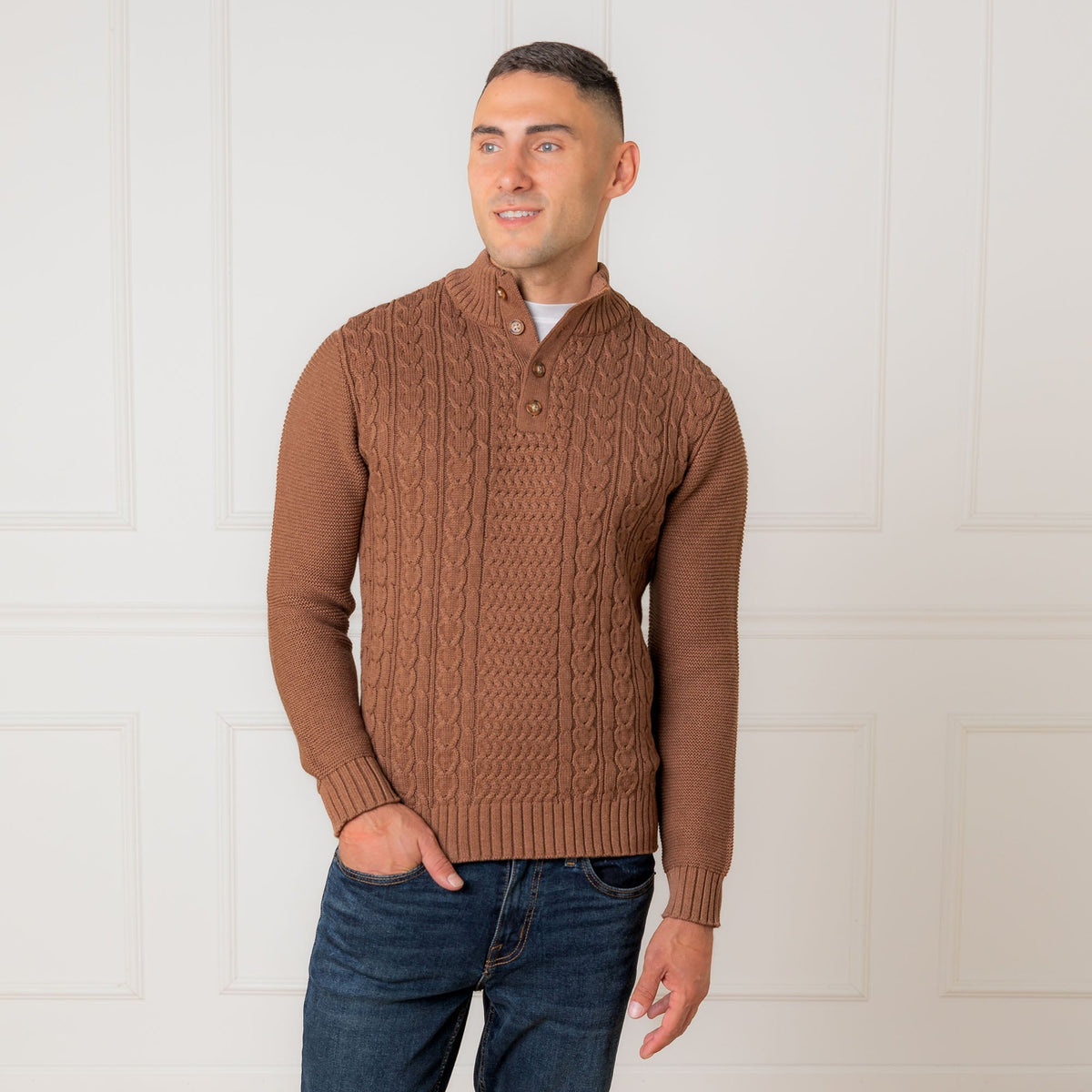 A man stands confidently wearing a cinnamon heather mock neck cable sweater made from organic cotton. The sweater features a textured cable design and a faux suede button placket.
