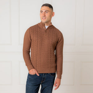 A man stands confidently wearing a cinnamon heather mock neck cable sweater made from organic cotton. The sweater features a textured cable design and a faux suede button placket.