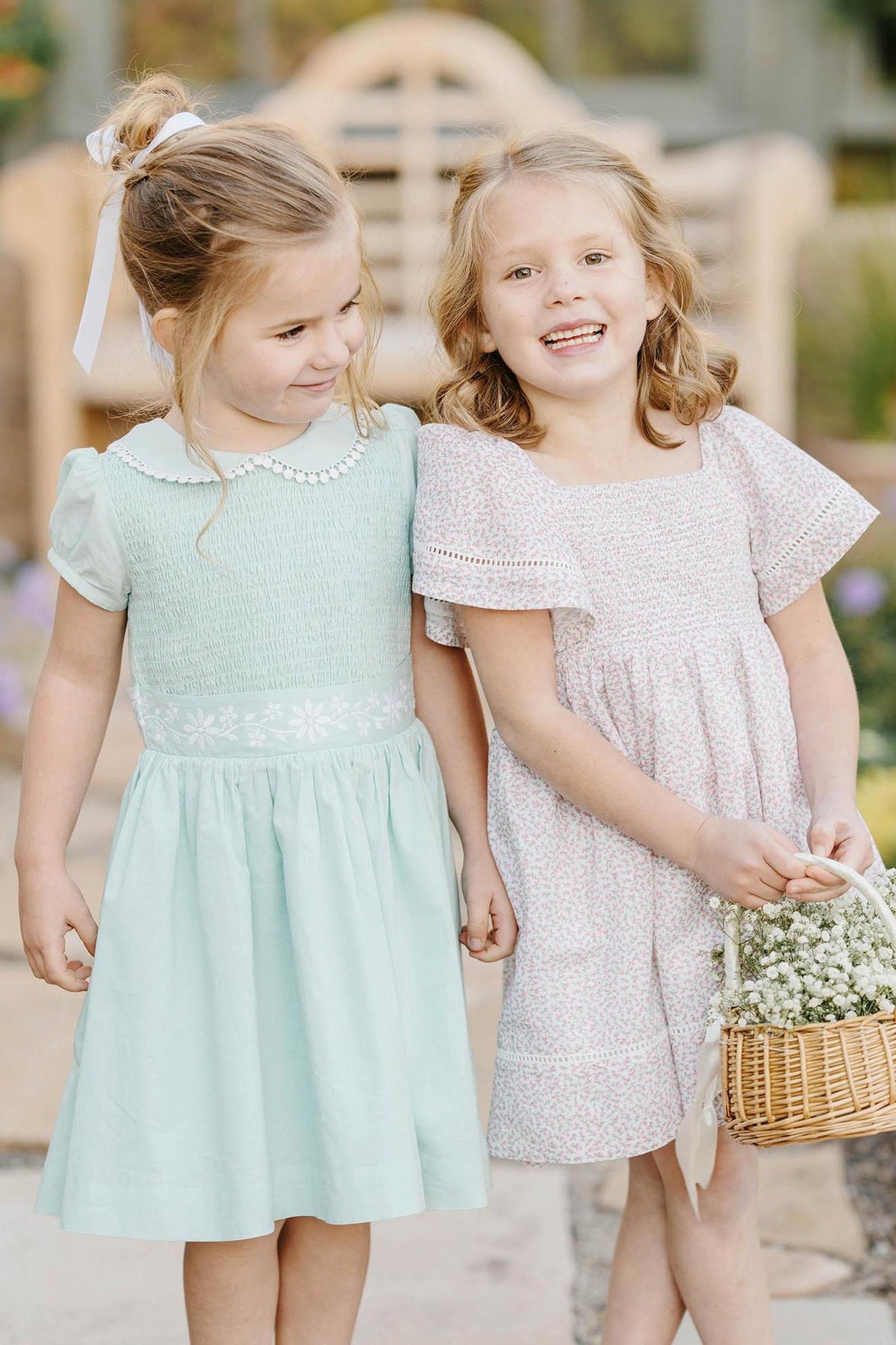 Two young girls in pastel dresses, one mint green with floral embroidery, the other in a floral print.
