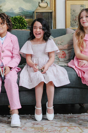 Three children sit on a couch, with the girl in the center wearing a white smocked dress with bell sleeves.