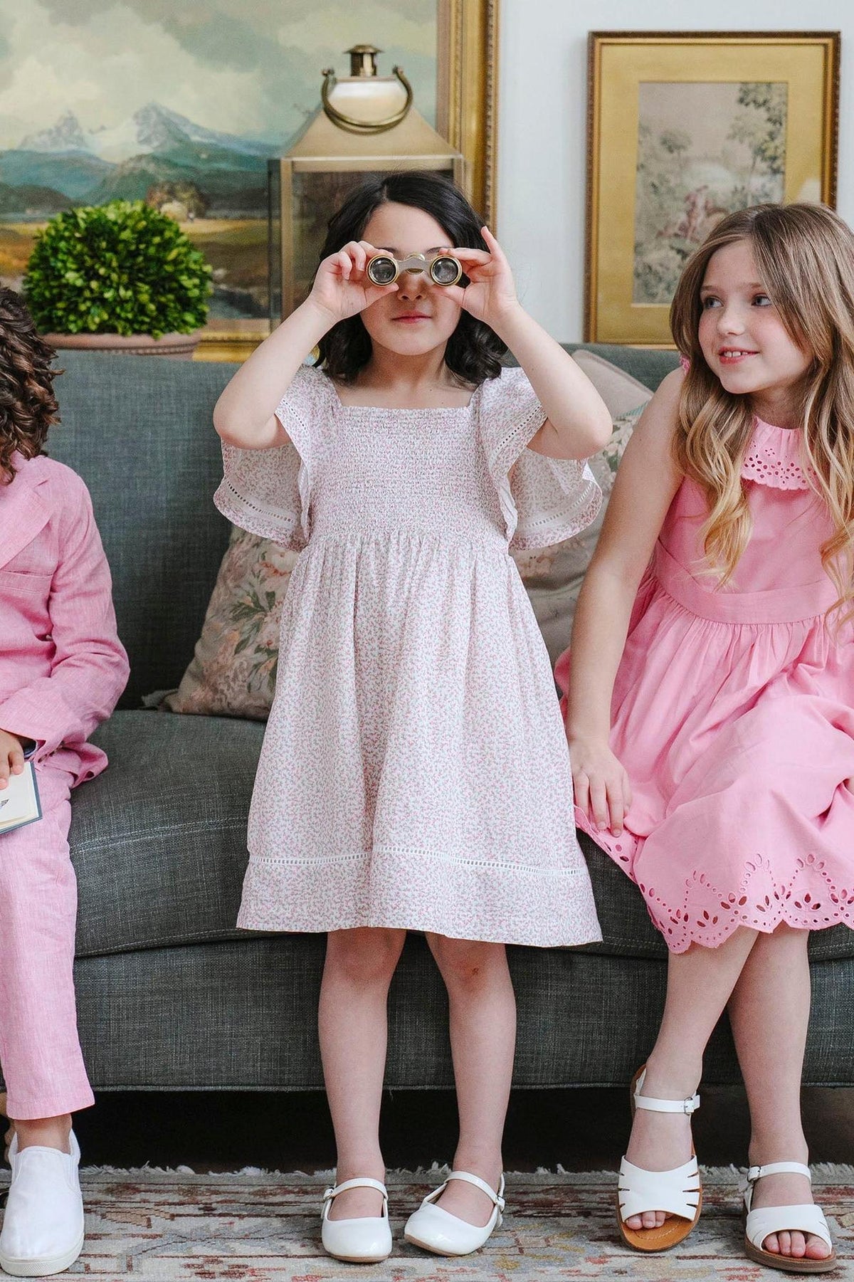 Three children sit on a couch; the girl in the center wears a floral smocked dress and holds binoculars.