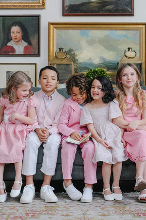 Five children in pastel pink and white clothing sit on a sofa, smiling in a room with framed paintings.