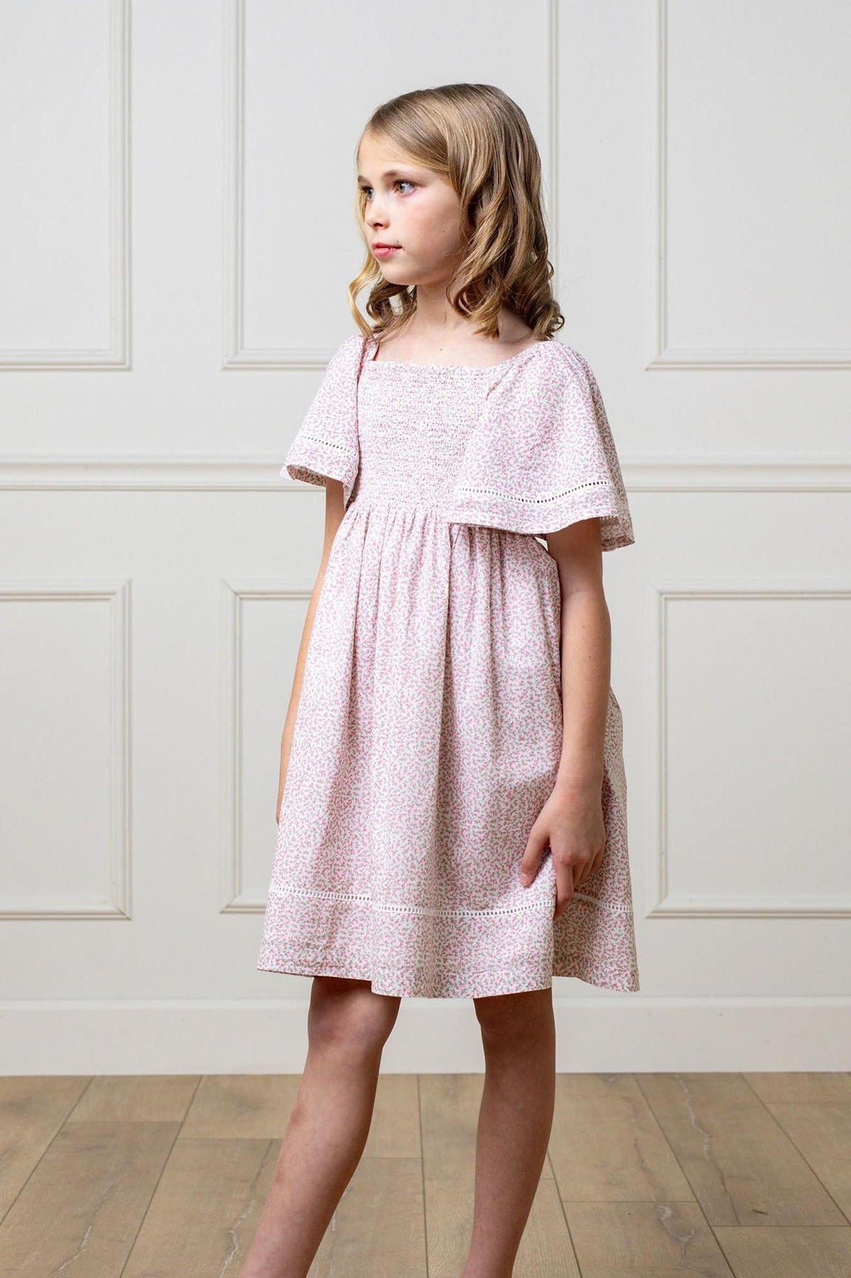 A young girl in a pink floral smocked dress with bell sleeves, standing in a room with paneled walls.