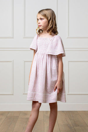 A young girl in a pink floral smocked dress with bell sleeves, standing in a room with paneled walls.