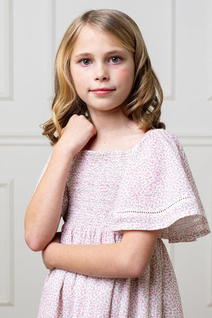 A young girl wears a pink floral smocked dress with bell sleeves, standing against a light background.