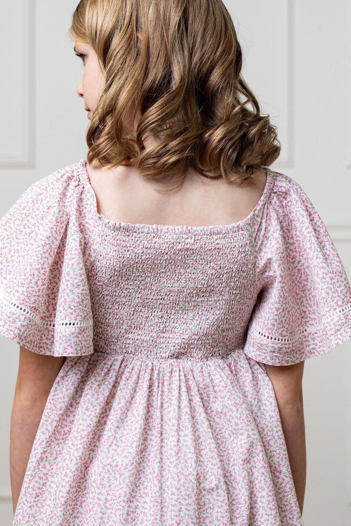 A young girl wearing a pink floral smocked dress with bell sleeves, showcasing a back view.