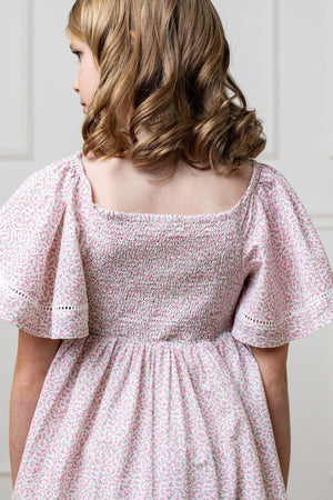 A young girl wearing a pink floral smocked dress with bell sleeves, showcasing a back view.
