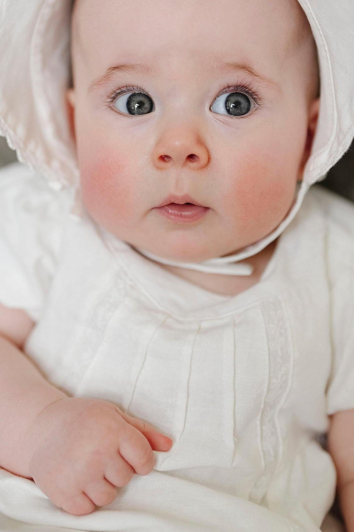 A close-up of a baby with big gray eyes, wearing a white linen dress with pintucking and a matching bonnet.