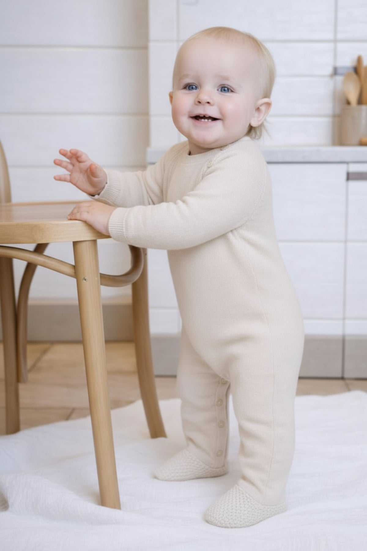 Baby in a oatmeal onesie standing next to a wooden chair in a kitchen.