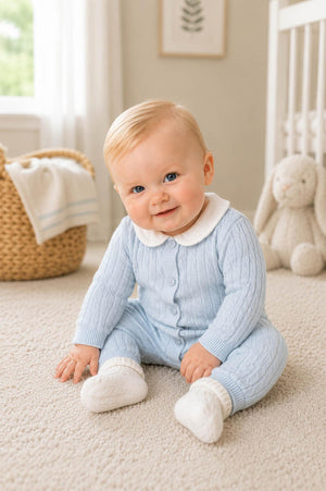 A baby sits on a soft carpet, wearing a light blue cable knit romper with a white Peter Pan collar. The background features a cozy.