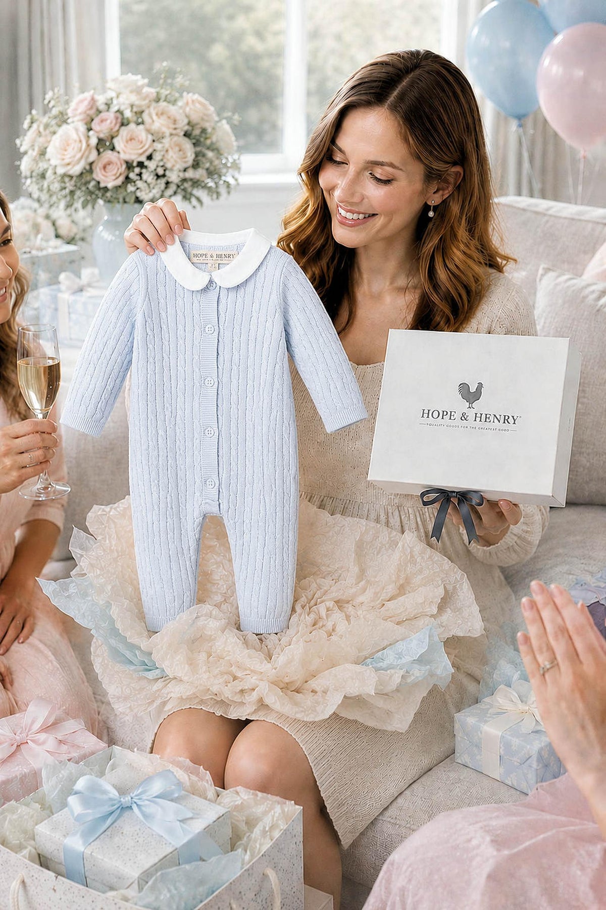 A woman holds a pale blue baby romper with a Peter Pan collar, surrounded by friends celebrating with champagne and gifts. The romper is made from soft, organic cotton.