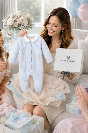 A woman holds a pale blue baby romper with a Peter Pan collar, surrounded by friends celebrating with champagne and gifts. The romper is made from soft, organic cotton.