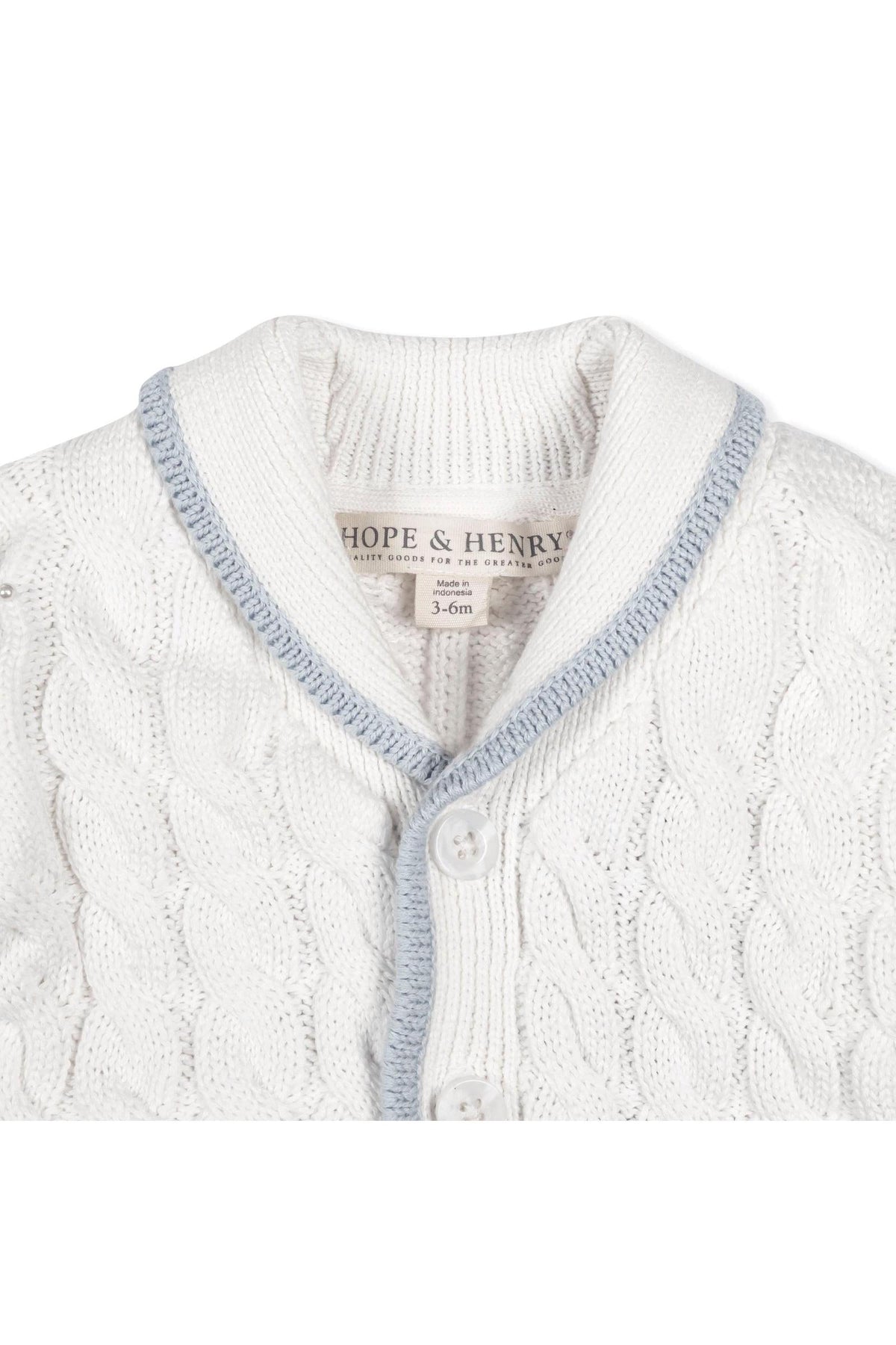Close-up of a white knitted cardigan with a light blue shawl collar, featuring a button closure and intricate cable knit design. Made from soft organic cotton for baby comfort.
