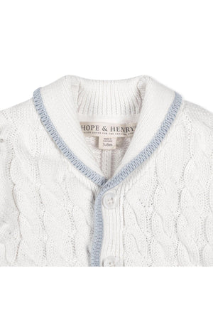 Close-up of a white knitted cardigan with a light blue shawl collar, featuring a button closure and intricate cable knit design. Made from soft organic cotton for baby comfort.