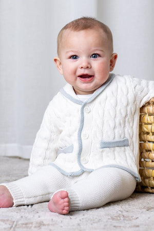 A smiling baby sits on a soft rug, wearing a creamy white cable-knit cardigan with gray trim and matching leggings, showcasing a cozy, stylish outfit perfect for everyday wear.
