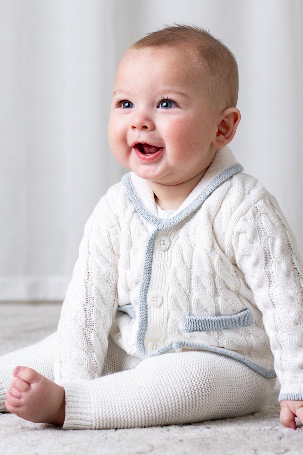 A smiling baby sits on a soft surface, wearing a cozy cream cardigan with a light blue collar and matching leggings, showcasing a classic and comfortable style.
