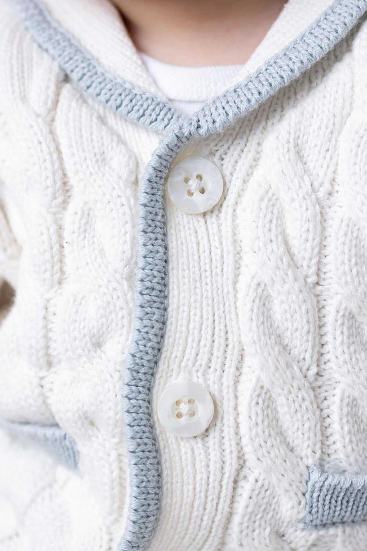 Close-up of a cream-colored knitted cardigan featuring a cable knit design, blue trim, and two large buttons. The soft organic cotton material ensures comfort for babies.