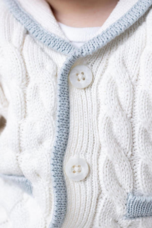 Close-up of a cream-colored knitted cardigan featuring a cable knit design, blue trim, and two large buttons. The soft organic cotton material ensures comfort for babies.