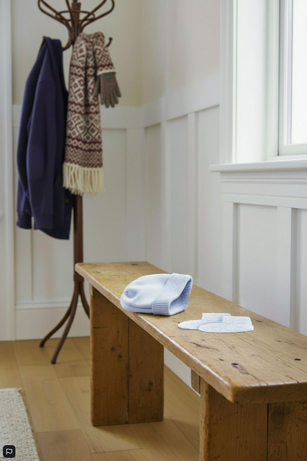 A pale blue knit beanie and matching booties are placed on a rustic wooden bench in a bright, cozy entryway.
