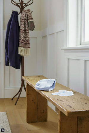 A pale blue knit beanie and matching booties are placed on a rustic wooden bench in a bright, cozy entryway.