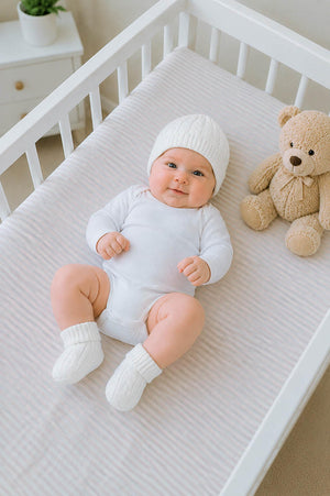 A smiling baby in a white onesie wears a cozy knitted white beanie and booties, lying on a soft, striped crib sheet beside a plush teddy bear.
