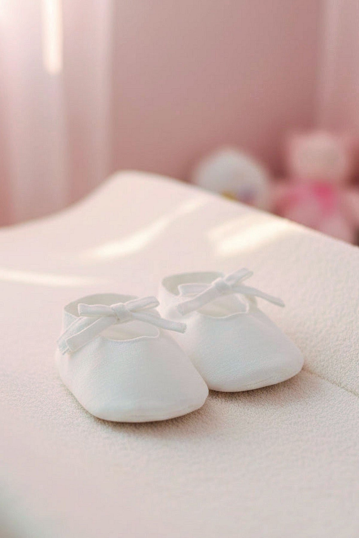 White linen baby shoes with decorative bows are placed on a soft, textured surface in a softly lit room.