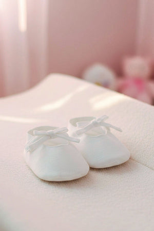 White linen baby shoes with decorative bows are placed on a soft, textured surface in a softly lit room.