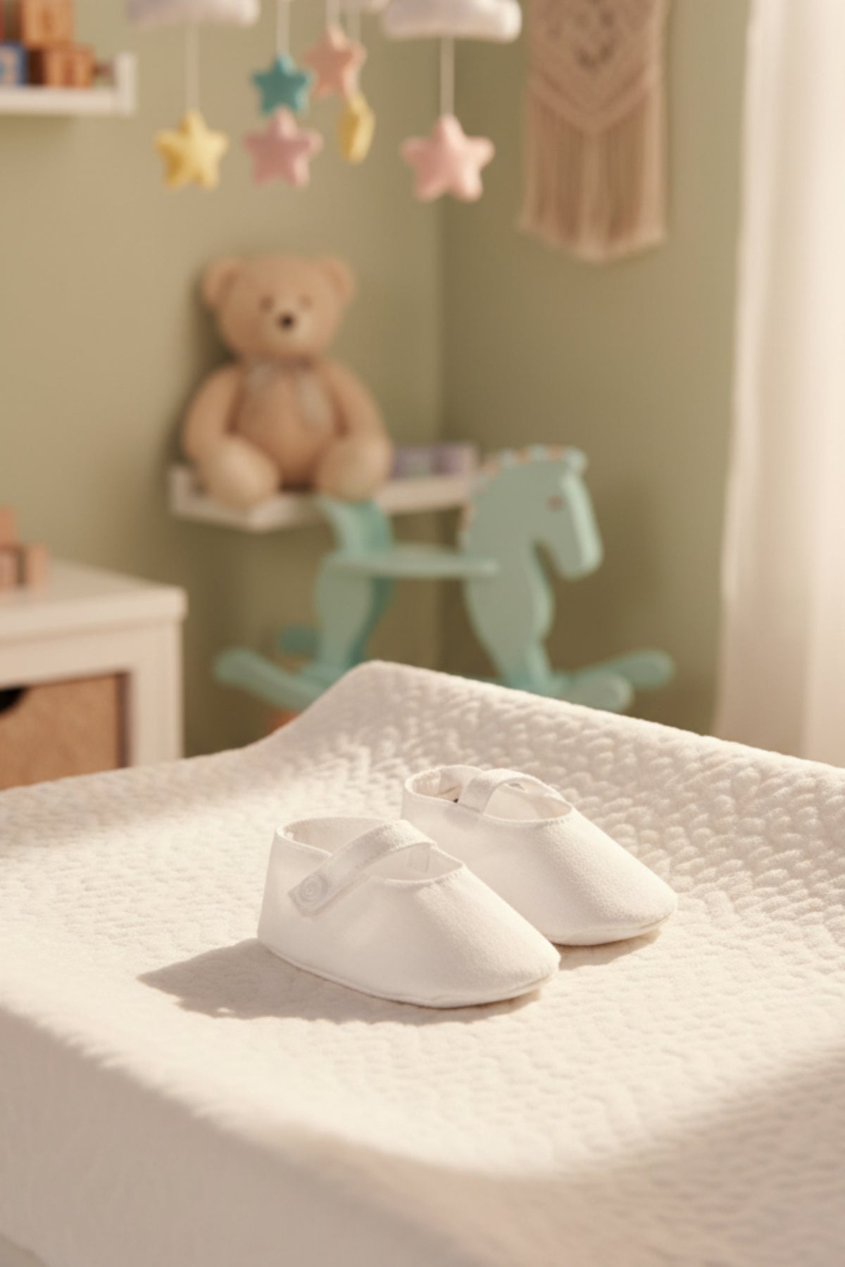 White linen baby shoes with a button strap are displayed on a textured blanket, set in a softly lit nursery.