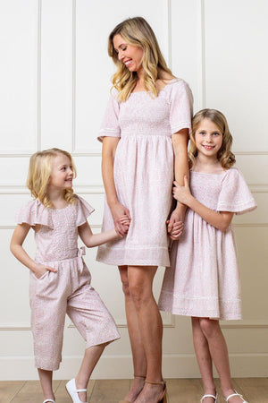 A woman and two girls wearing matching light pink floral outfits, smiling in a room with white paneled walls.