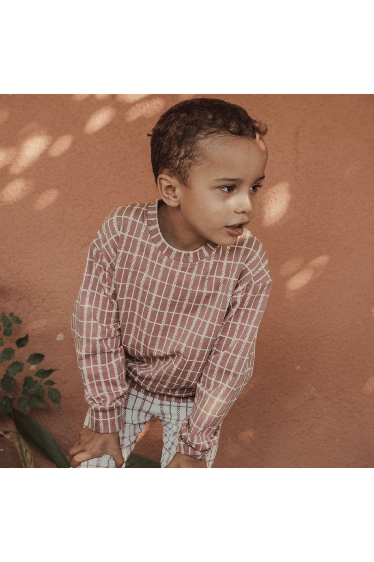 A child wearing a mauve check printed cotton sweatshirt and matching leggings poses against a warm, textured wall. The outfit features a soft, comfortable fabric ideal for casual wear.