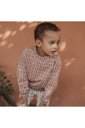 A young child wearing a mauve check printed cotton sweatshirt stands against a warm, textured wall. The sweatshirt features a relaxed fit, perfect for cozy outings or lounging.