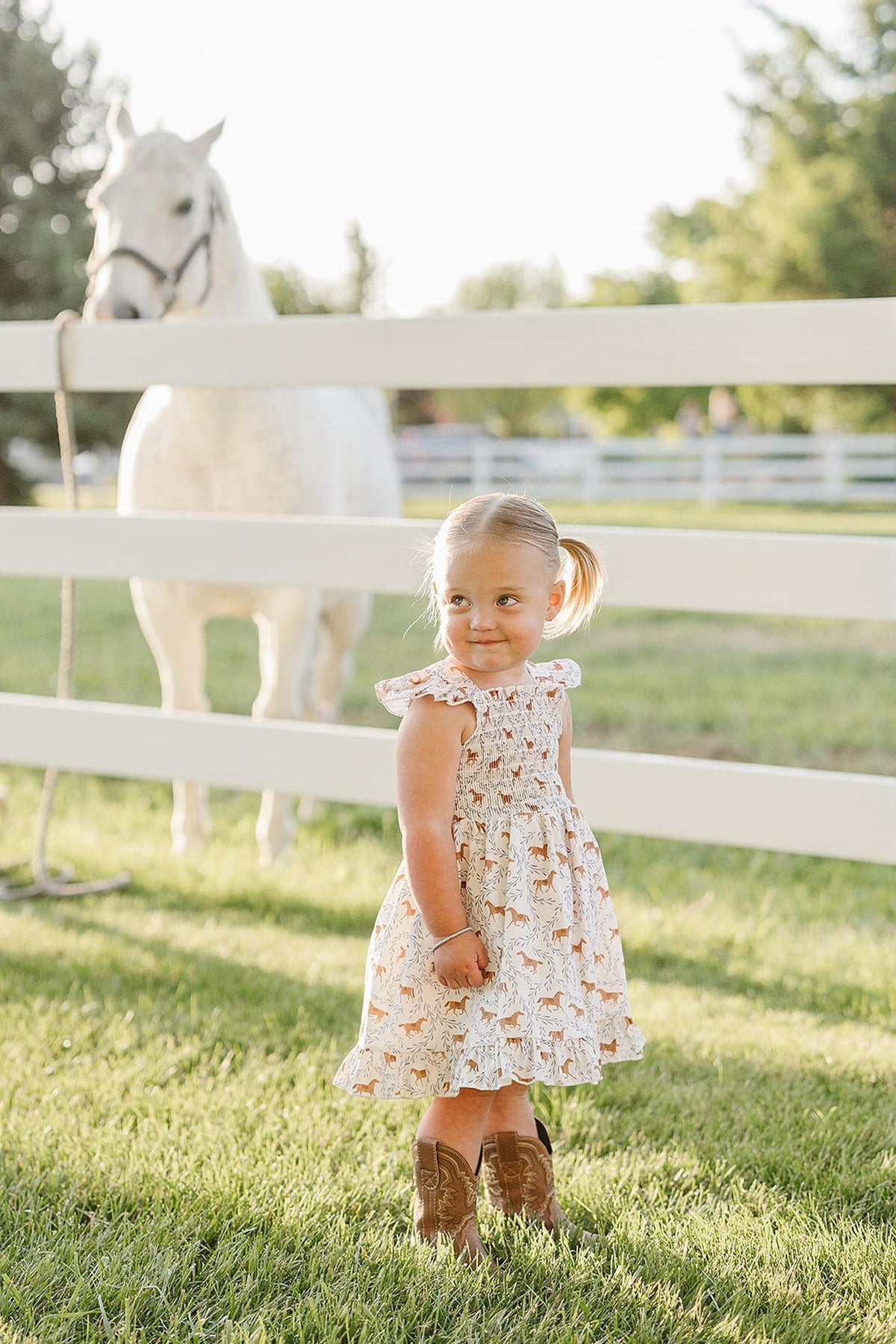 A young girl wearing a pony-printed smocked dress stands in a grassy field near a white horse, surrounded by a white fence and green trees in the background.