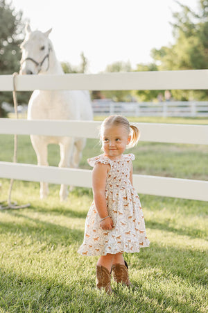 A young girl wearing a pony-printed smocked dress stands in a grassy field near a white horse, surrounded by a white fence and green trees in the background.