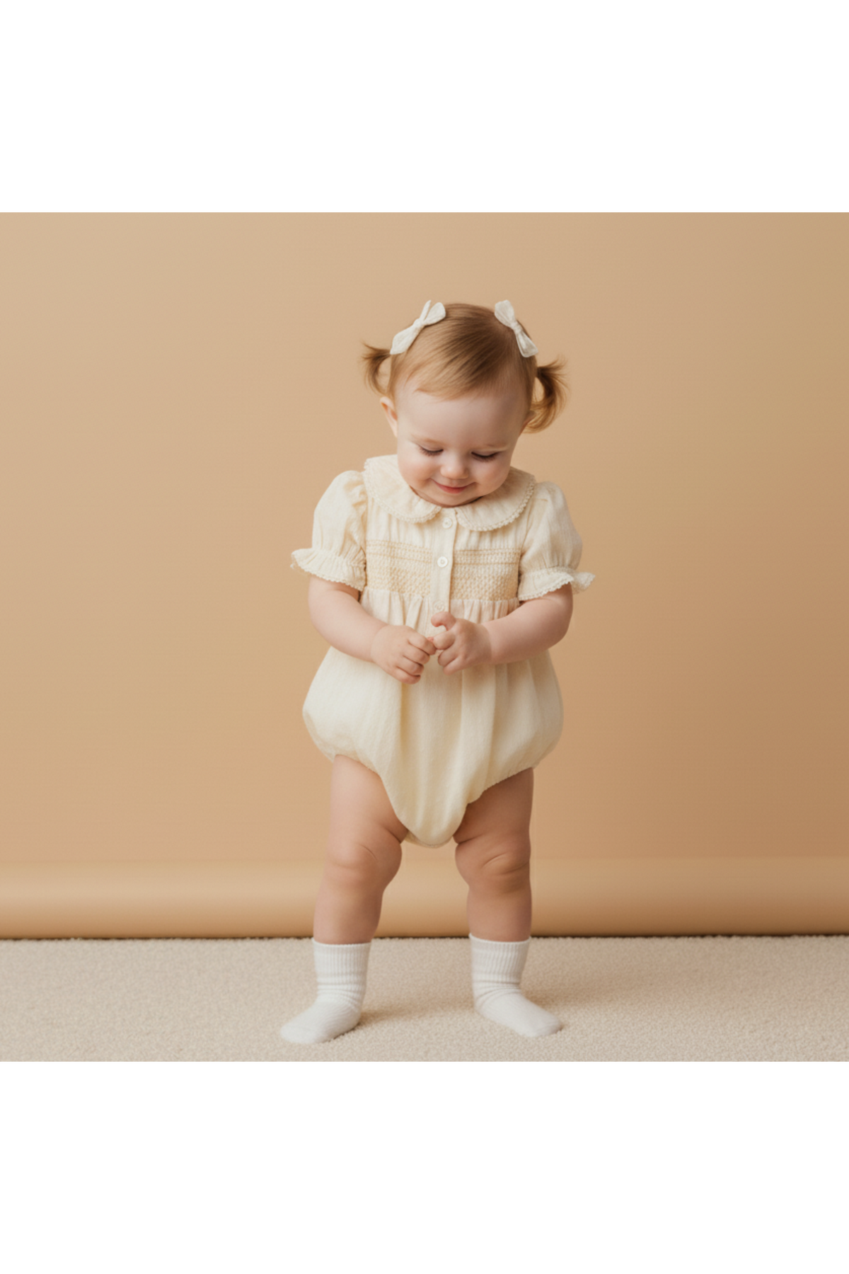 A happy baby wearing a butter yellow organic cotton smocked romper with short ruffled sleeves stands on a soft surface, smiling and playing, against a warm, neutral backdrop.