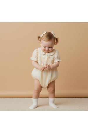 A happy baby wearing a butter yellow organic cotton smocked romper with short ruffled sleeves stands on a soft surface, smiling and playing, against a warm, neutral backdrop.