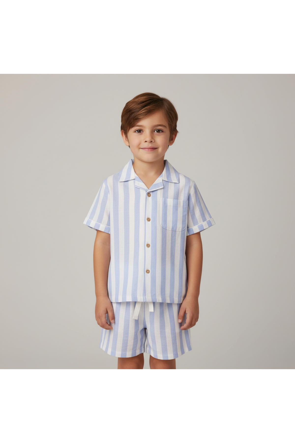 A young boy smiles while wearing a light blue and white striped organic cotton shirt and shorts set. The outfit features a button-down shirt with a pocket, perfect for summer.