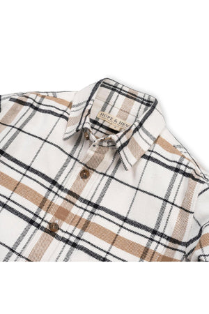 A soft white plaid shirt featuring brown and black stripes, made from organic cotton. The shirt is designed for boys and showcases a classic, timeless style suitable for various occasions.