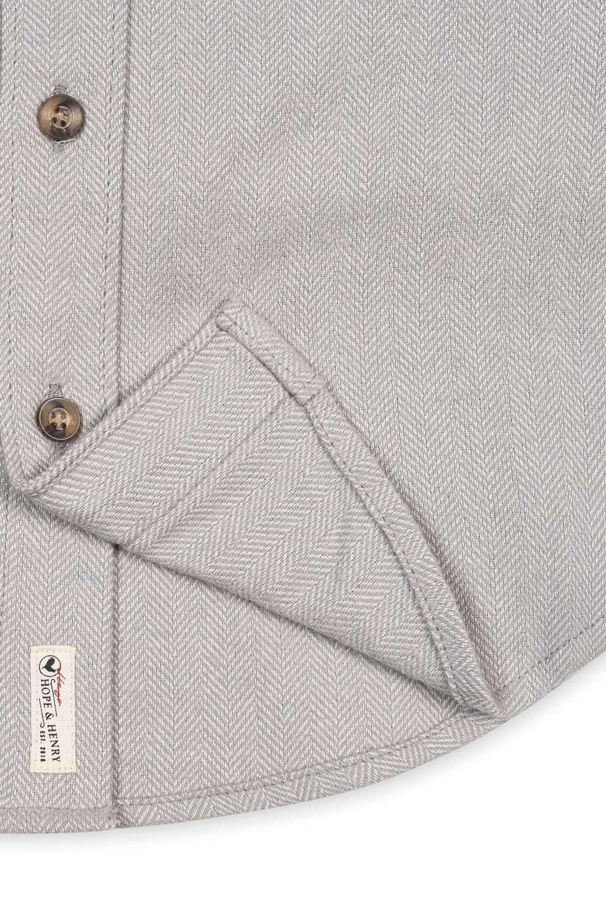 Close-up of a grey organic cotton flannel shirt featuring a herringbone pattern, button details, and a tag indicating sustainable materials. The fabric appears soft and textured, ideal for all ages.