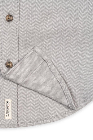 Close-up of a grey organic cotton flannel shirt featuring a herringbone pattern, button details, and a tag indicating sustainable materials. The fabric appears soft and textured, ideal for all ages.