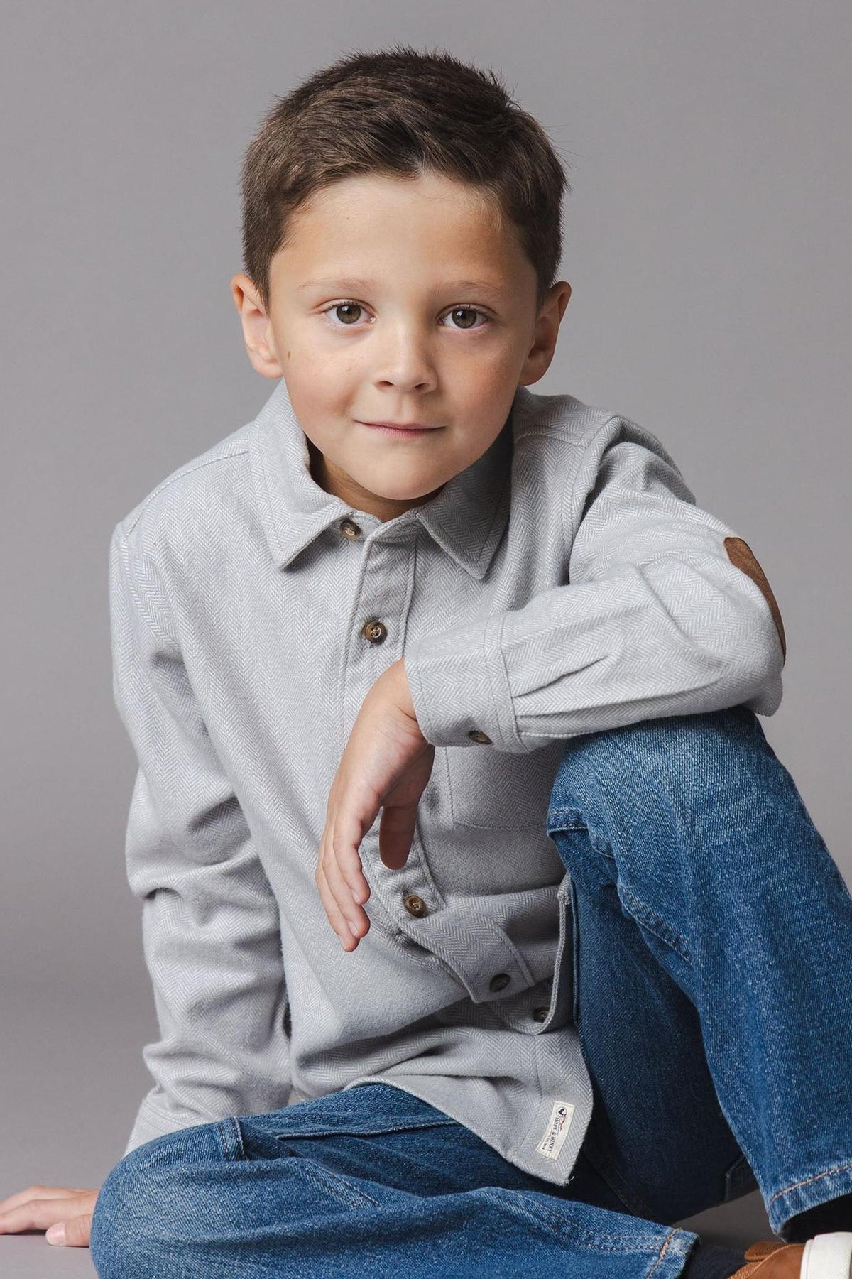 A young boy sits casually, wearing a light grey organic cotton flannel shirt with buttons and elbow patches, paired with blue jeans against a neutral grey background.