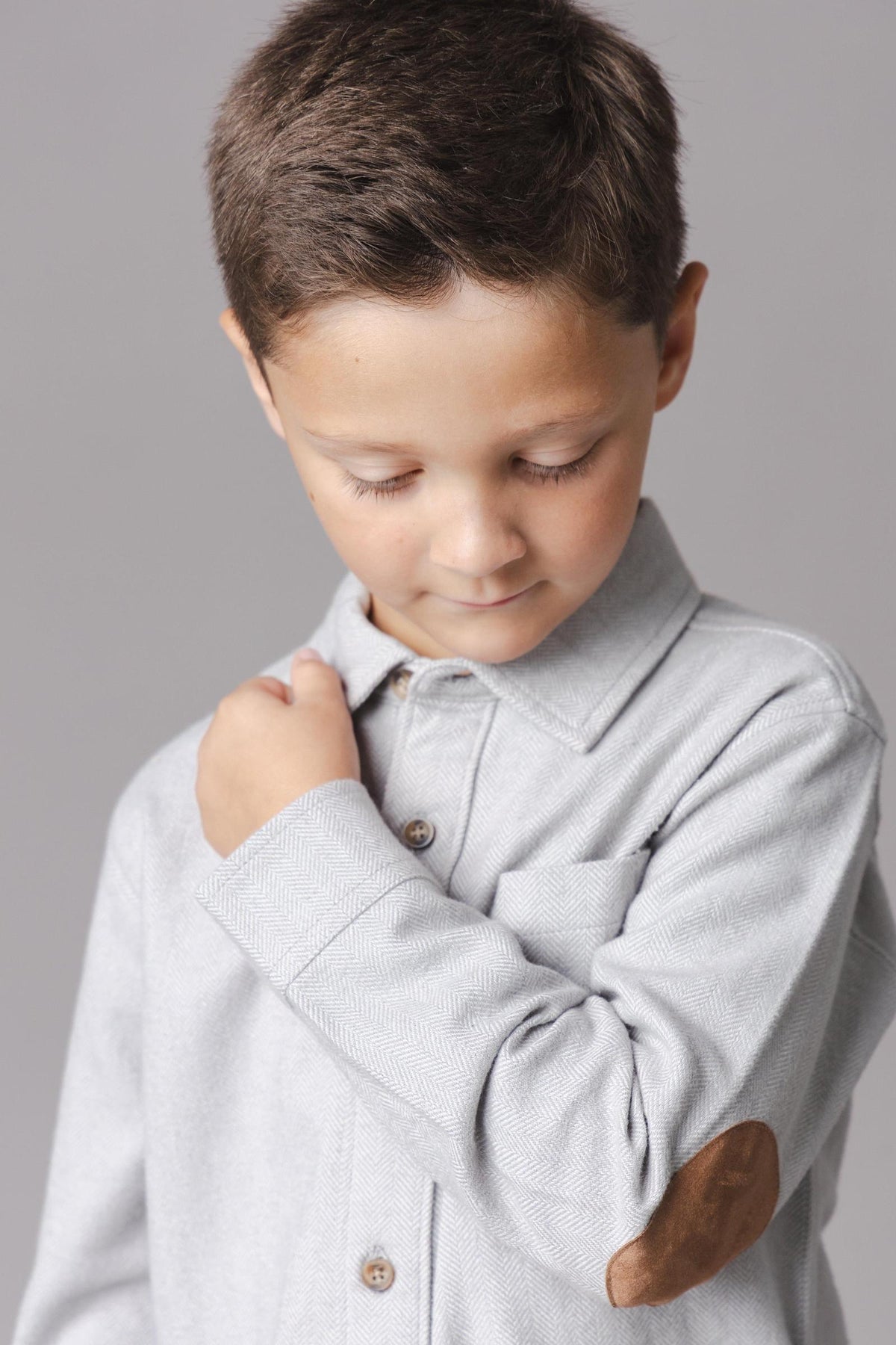 A young boy is wearing a light grey organic cotton flannel shirt with a subtle herringbone pattern and a brown elbow patch.