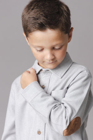 A young boy is wearing a light grey organic cotton flannel shirt with a subtle herringbone pattern and a brown elbow patch.