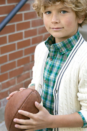 A young boy with curly hair holds a brown football, wearing a green plaid shirt under a cream cable-knit cardigan. The background features a brick wall, adding a rustic touch.