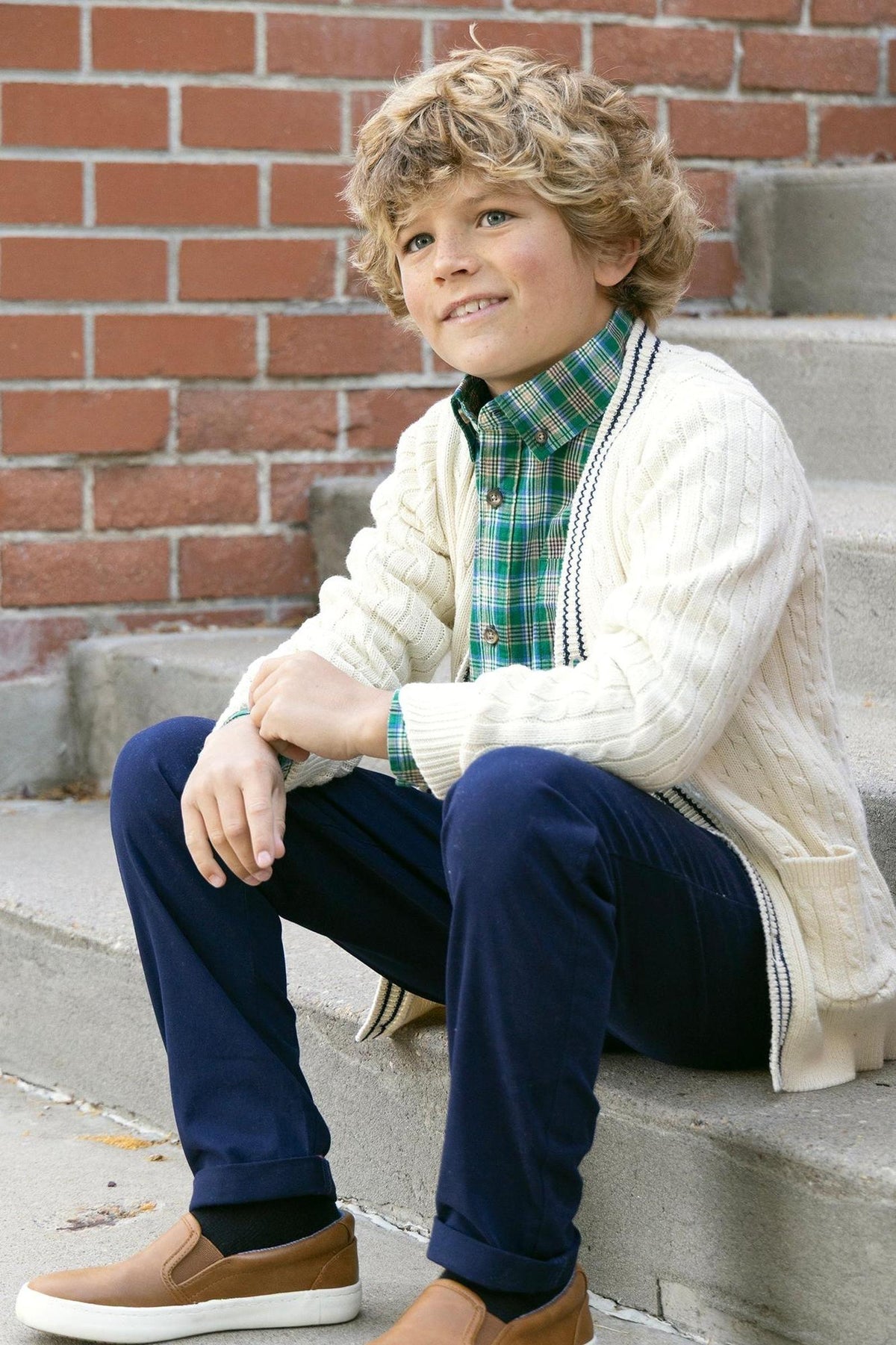 A boy with curly hair sits on stone steps, wearing a green classic plaid shirt under a cream cable-knit cardigan, paired with navy pants and brown slip-on shoes.