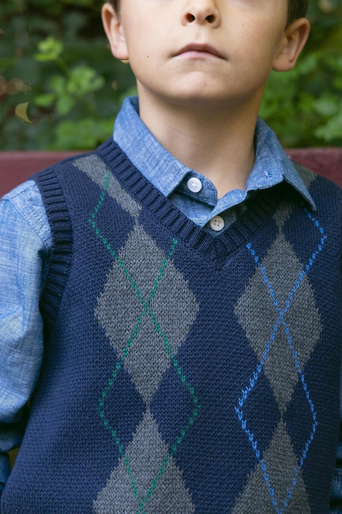 A young boy models a navy and gray argyle sweater vest made from soft organic cotton, layered over a light blue shirt, showcasing a classic, polished look.