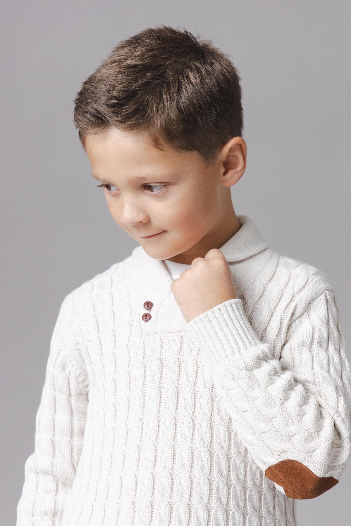 A young boy models a light oatmeal organic cotton shawl collar cable sweater, featuring a cozy design and button closure.